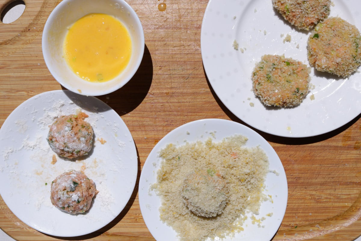 Two salmon balls dusted in flour, bowl of beaten egg, plate with salmon ball coated in panko, plate with 3 breaded salmon balls.