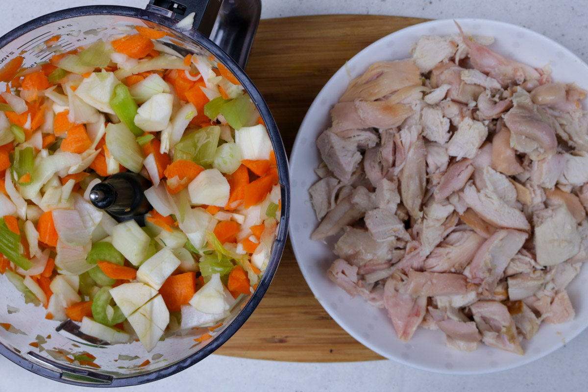 Chopped veggies in a food processor bowl and diced cooked chicken in another bowl.